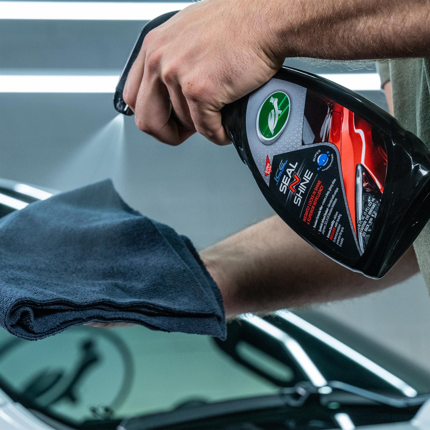 Turtle Wax ICE Seal N Shine being sprayed on a car windshield, alongside a microfiber cloth. Car care and detailing product for vehicles.