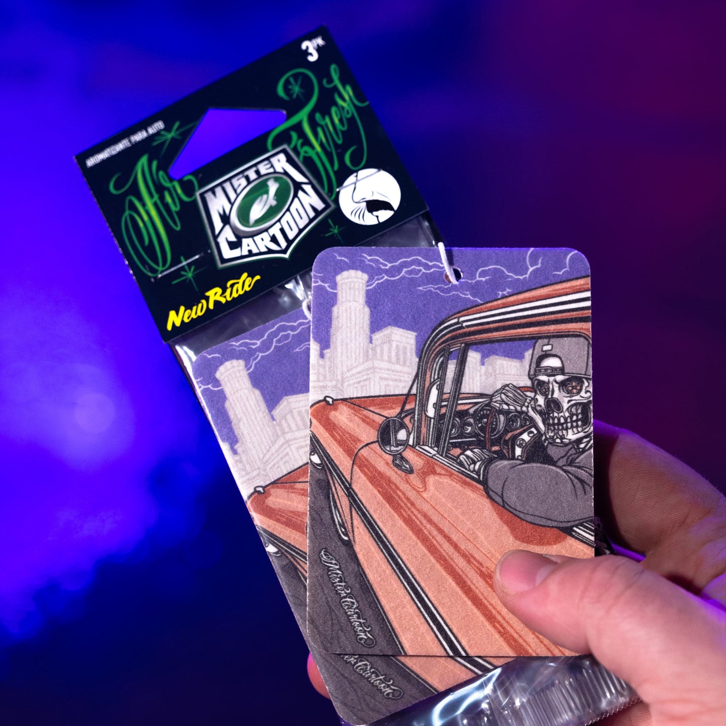 Auto air freshener with a "New Ride" scent. Cartoon skull driving a lowrider car in front of city buildings. Mister Cartoon design.
