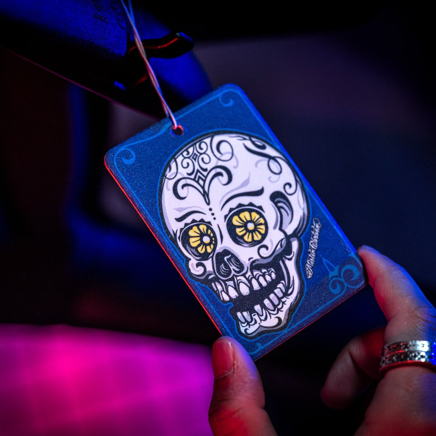 Here's a description of the image in under 125 characters:

Car air freshener with a blue frame and skull design (sugar skull art) hanging in auto. "Midnight" vibe.
