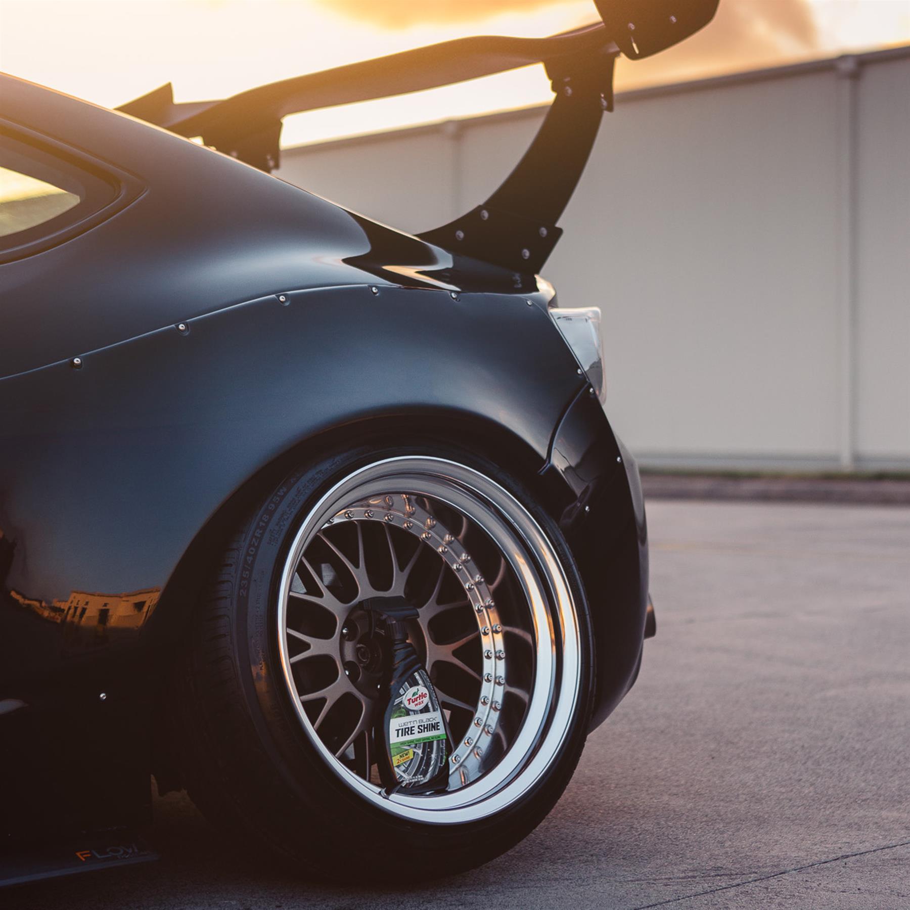 Black car with spoiler features shiny tires. Turtle Wax Tire Shine bottle is visible inside the wheel.