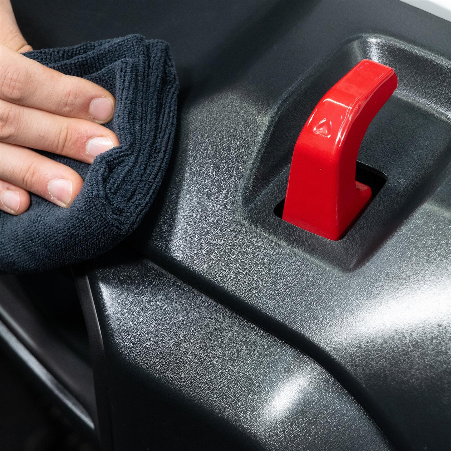 Detailing auto interior trim. Microfiber cloth wiping textured grey surface near red handle.