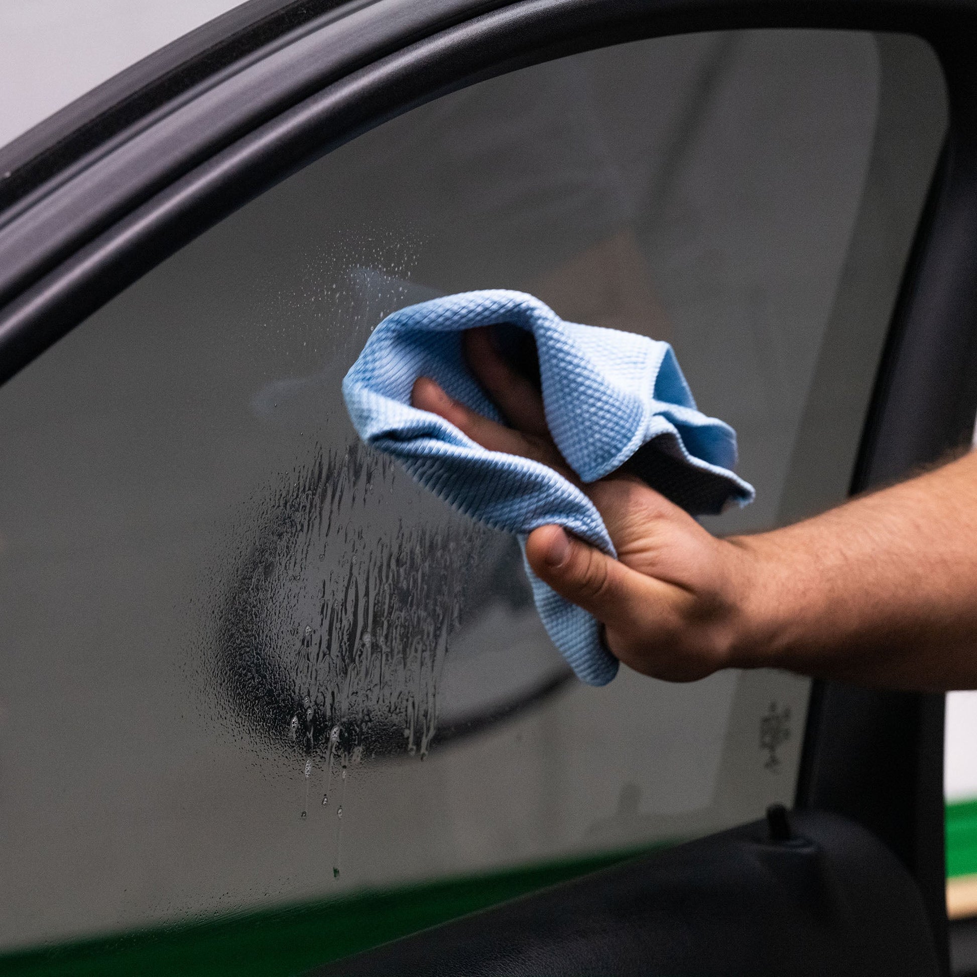 Hand wiping a tinted car window with a blue cloth. ClearVue residue visible. Automotive detailing for clean visibility, India.