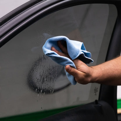 Hand wiping a tinted car window with a blue cloth. ClearVue residue visible. Automotive detailing for clean visibility, India.